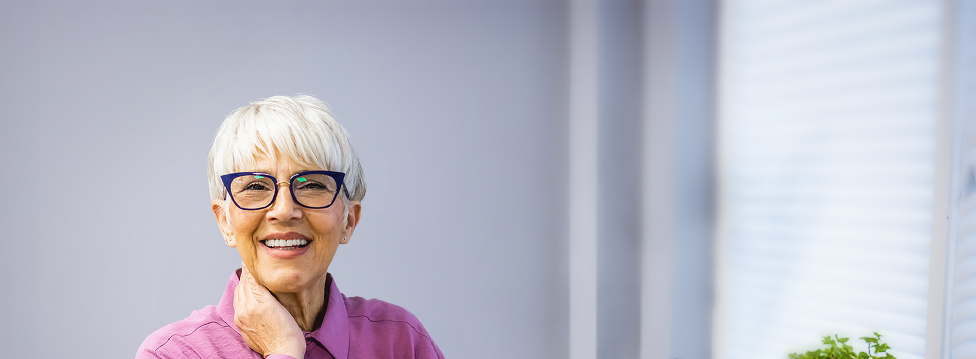 The image depicts an older woman with short hair wearing glasses, a pink top, and a blue cardigan, seated at a table with a plant in front of her, smiling and looking directly at the camera.