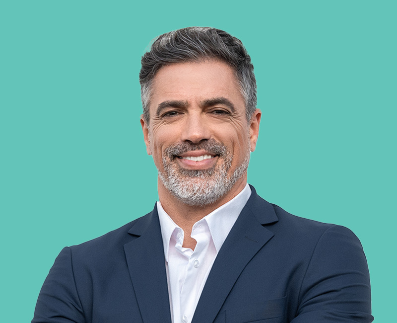 This image features a man with gray hair, wearing a suit jacket over a blue shirt, smiling at the camera against a light green background.